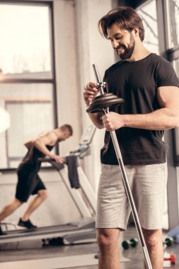 happy handsome sportsman fixing weights on iron bar for training in gym clipart