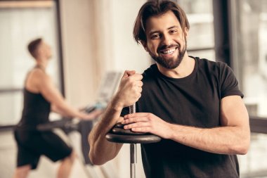 smiling sportsman leaning on iron bar with weight plates in gym clipart