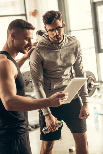 handsome trainer showing results of exercising on tablet to exhausted sportsman in gym