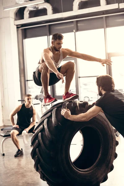 Sportovní Mladý Muž Skákání Pneumatiky Při Tréninku Přáteli Tělocvičně — Stock fotografie