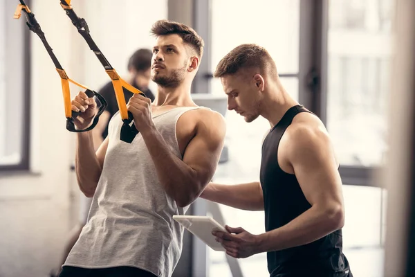 Trainer Using Digital Tablet Helping Muscular Sportsman Exercising Fitness Straps — Stock Photo, Image