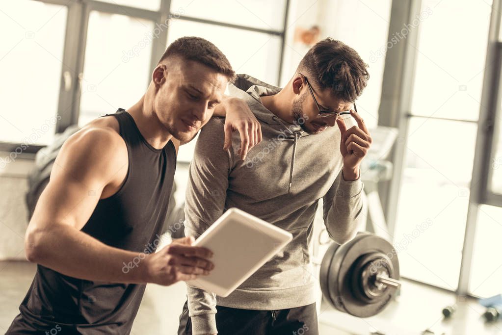 handsome trainer showing results of exercising on tablet to tired sportsman in gym