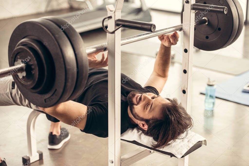 smiling handsome sportsman preparing lifting barbell on bench press in gym