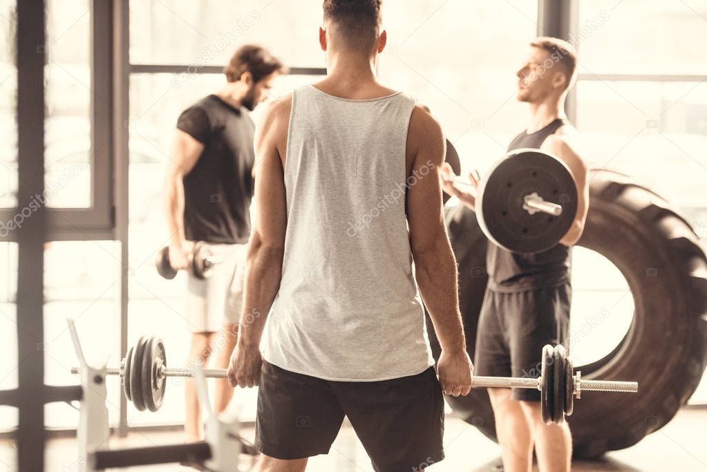 back view of muscular young man lifting barbell and looking at friends in gym