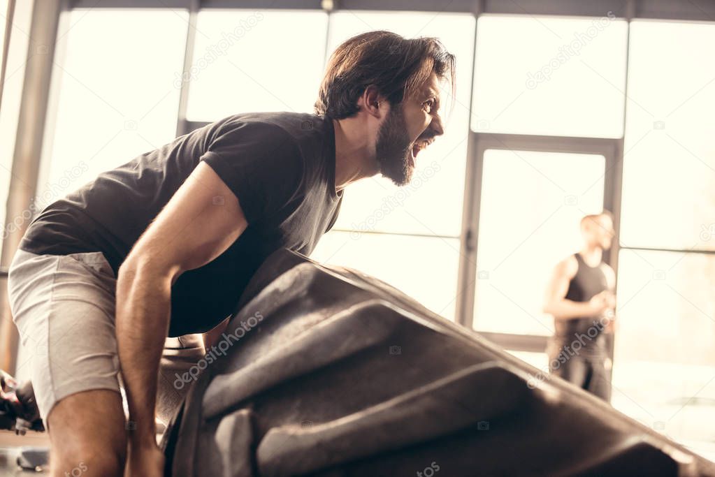 side view of athletic man lifting tire and screaming in gym 