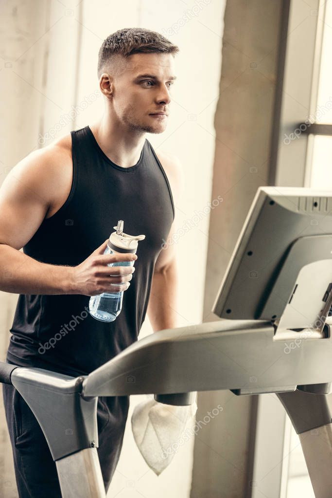 Sporty young man holding bottle of water and training on treadmill