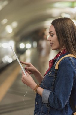 kulaklık ve metro istasyonunda dijital tablet ile gülümseyen kadın yan görünüm