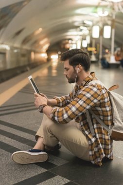 Dijital tablet ve metro istasyonunda katta oturan kulaklıklar ile şık adam yan görünüm 
