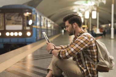 Dijital tablet ve metro istasyonunda katta oturan kulaklıklar genç adamla yan görünüm 