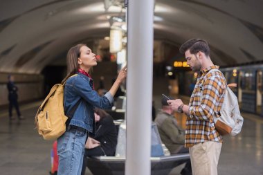 kadın Gezgin bilgi yönetim kurulu ve metro istasyonunda smartphone kullanarak erkek sırt çantası ile yan görünüm 