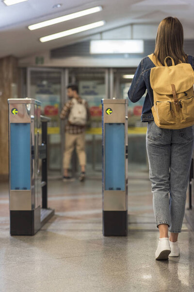 rear view of female stylish traveler with backpack passing through turnstiles and male tourist behind