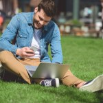 Man with laptop looking at smartphone screen and sitting on grass