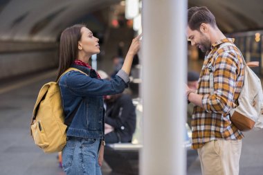 kadın Gezgin bilgi yönetim kurulu ve metro istasyonunda smartphone kullanarak erkek bakarak yan görünüm 