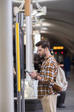 sırt çantası ve Metro İstasyonu'na bilgi tahtaya bakarak smartphone ile genç erkek turist yan görünüm 