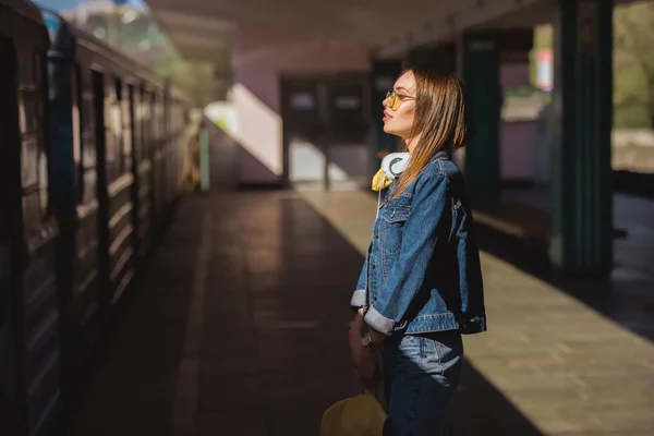 屋外の地下鉄駅で立っているヘッドフォンとサングラスでスタイリッシュな女性の側面図 — ストック写真