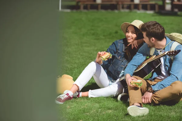 ギターと草の上に座ってサンドイッチの帽子で観光客のカップルの笑顔  — 無料ストックフォト