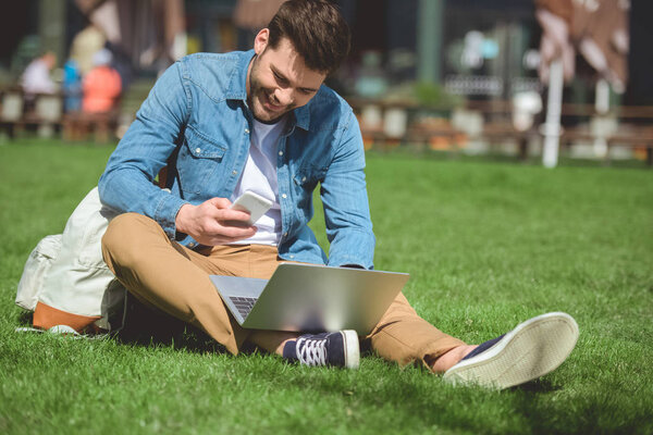 man with laptop looking at smartphone screen and sitting on grass