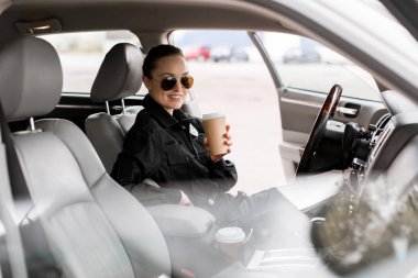 smiling policewoman with paper cup of coffee sitting in car and looking at camera clipart