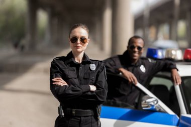 young serious policewoman standing with crossed arms while her partner standing near car and smiling blurred on background clipart
