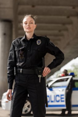 attractive female police officer in uniform looking at camera with blurred partner near car on background clipart