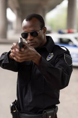 young african american policeman in sunglasses aiming by handgun  clipart