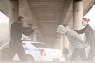 side view of angry policeman aiming handgun on male criminal while partner holding him  clipart