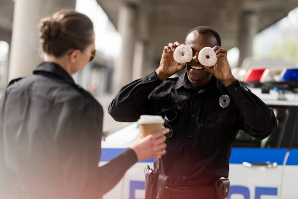 african american police officer pretending doughnuts as his eyes to amuse his partner