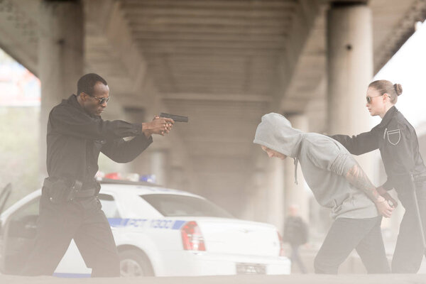 side view of angry policeman aiming handgun on male criminal while partner holding him 