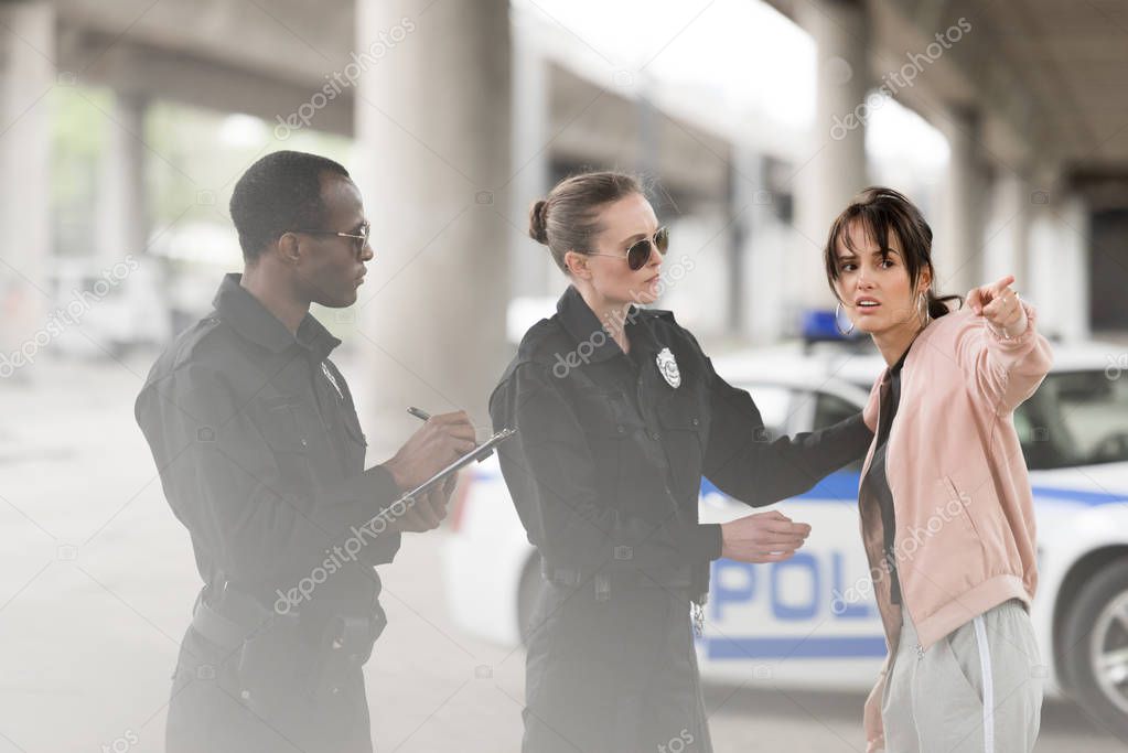 young worried woman pointing by finger to policewoman and policeman writing in clipboard 
