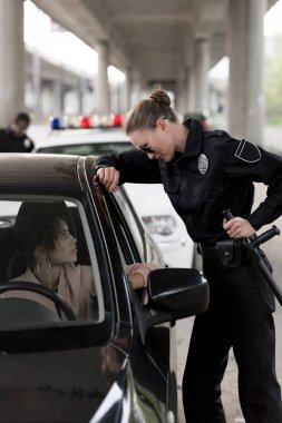 side view of policewoman holding truncheon and talking to young woman sitting in car  clipart