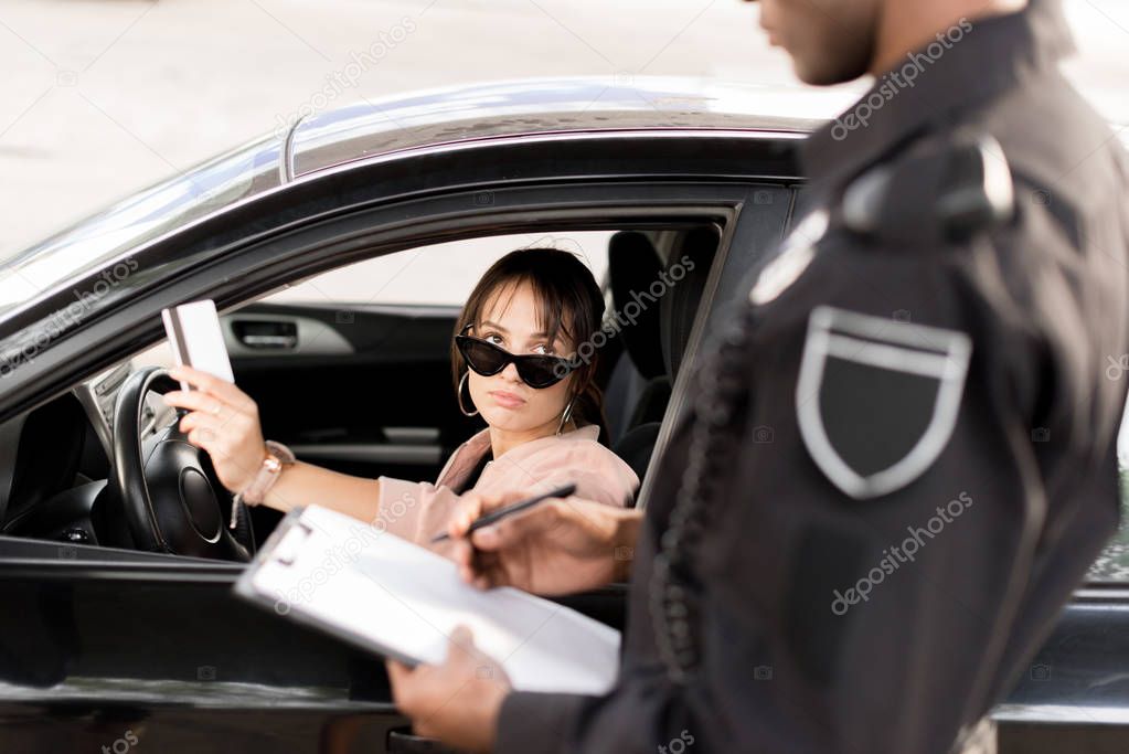 Cropped image of policeman with clipboard and pen talking to young driver in car giving driver license