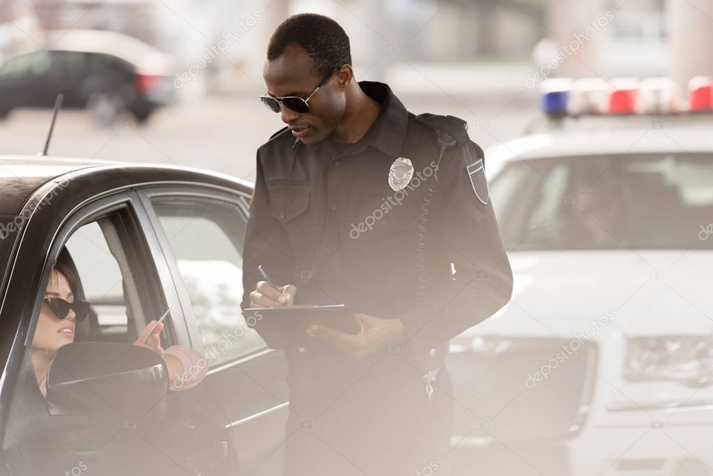 young woman in car giving driver license to african american policeman with clipboard and pen