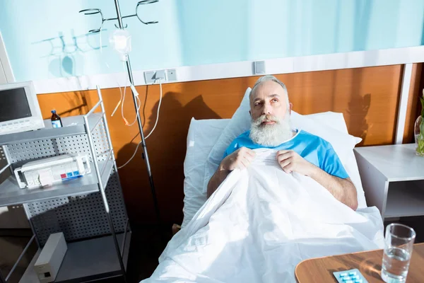 Sick senior man lying in hospital bed and adjusting blanket — Stock Photo