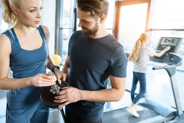 Jovem homem e mulher de pé com barbell enquanto pequeno treino menina na esteira no ginásio — Fotografia de Stock