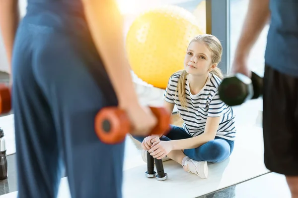 Gente de fitness haciendo ejercicio con pesas mientras una adolescente se sienta en la alfombra en el gimnasio - foto de stock