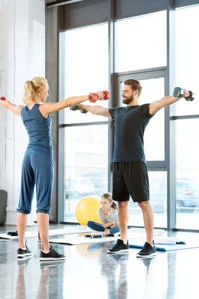 Gente de fitness haciendo ejercicio con pesas mientras una adolescente se sienta en la alfombra en el gimnasio - foto de stock