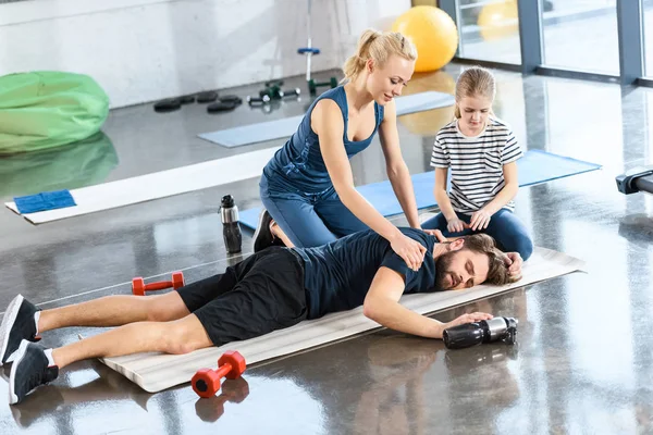 Mulher com menina ajudando o homem cansado deitado no tapete no ginásio — Fotografia de Stock