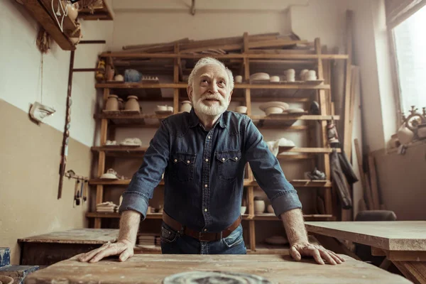 Front view of senior potter standing and leaning on table against shelves with pottery goods at workshop — Stock Photo