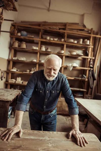 Front view of senior potter standing and leaning on table against shelves with pottery goods at workshop — Stock Photo