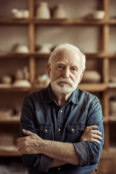 Ritratto di ceramista anziano in piedi contro scaffali con ceramiche in officina — Foto stock