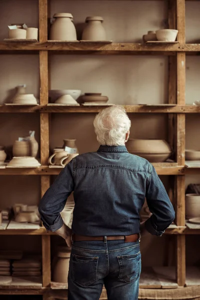 Vista trasera del alfarero senior parado cerca de estantes con artículos de cerámica y buscando algo en el taller — Stock Photo