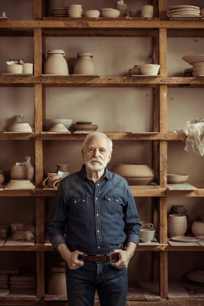 Vista anteriore del vasaio anziano in piedi contro scaffali con ceramiche in officina — Foto stock