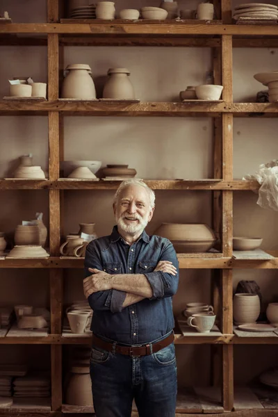 Front view of senior potter standing against shelves with pottery goods at workshop — Stock Photo