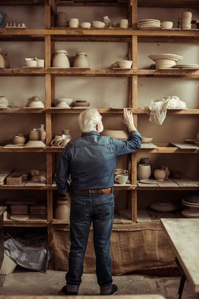 Vista posteriore del vasaio anziano in piedi vicino a scaffali con ceramiche e alla ricerca di qualcosa in officina — Foto stock