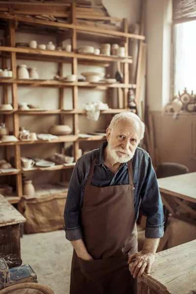 Front view of senior potter in apron standing and leaning on table against shelves with pottery goods at workshop — Stock Photo