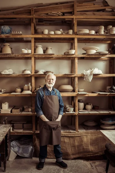 Vue de face du potier senior dans le tablier debout contre des étagères avec des articles de poterie à l'atelier — Photo de stock