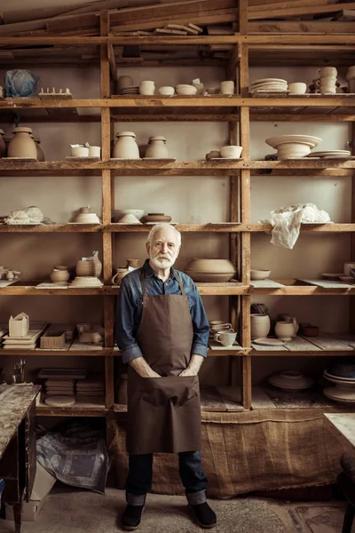 Vue de face du potier senior dans le tablier debout contre des étagères avec des articles de poterie à l'atelier — Photo de stock