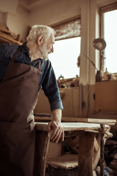 Seitenansicht des Senior-Töpfers in Schürze stehend und auf einem Tisch in der Werkstatt angelehnt — Stockfoto