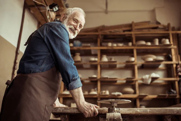 Side view of senior potter in apron standing and leaning on table against shelves with pottery goods at workshop — Stock Photo