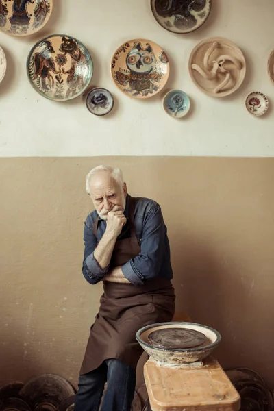 Alfarero senior en delantal pensando y de pie contra la pared con artículos de cerámica colgantes - foto de stock
